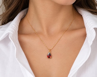 May include: A gold chain necklace featuring a single teardrop-shaped red gemstone pendant. The pendant is set in a gold metal frame and hangs from a delicate gold link chain, resting against a white collared shirt.