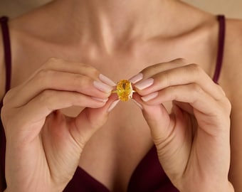 May include: A gold ring featuring a large, oval-cut yellow gemstone. The ring is held up in front of a person, showcasing the bright, eye-catching stone. The ring's band is a simple, elegant design.