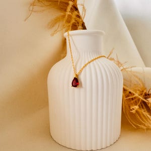 May include: A gold-colored necklace with a teardrop-shaped, deep red gemstone pendant. The necklace is displayed on a white, ribbed vase, with dried ornamental grass in the background. The pendant is approximately 0.75 inches in length.