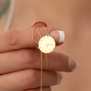 May include: A gold-coloured necklace with a circular pendant designed to resemble a clock face. The pendant has a delicate chain and displays the numbers 1 to 12 around the edge, with hands indicating the time. The necklace is a jewellery item.