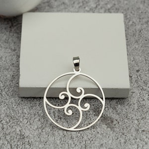 May include: A silver pendant with a circular design featuring swirling, scroll-like patterns. The pendant has a small loop at the top for a chain. The design is open, allowing light to pass through. The pendant is displayed on a gray surface.