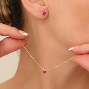 Pode incluir: Um delicado colar de corrente de ouro com um pequeno pingente de pedra vermelha. Brincos de pressão de pedra vermelha combinando também são mostrados.