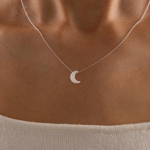 Puede incluir: Un delicado collar de plata con un colgante de media luna. El dije de luna es pequeño y está centrado en una fina cadena. El collar se luce sobre una camiseta clara, resaltando la sencilla elegancia de la joya.