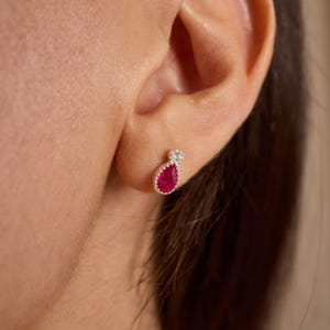 May include: A close-up of a silver earring with a teardrop-shaped, bright red gemstone. The earring has a small, round, clear stone at the top. The earring is worn on an ear.
