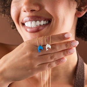 May include: Two dolphin pendant necklaces. One pendant is blue, the other is white. Both pendants are on delicate gold chains. The necklaces are held up in front of a person's face.