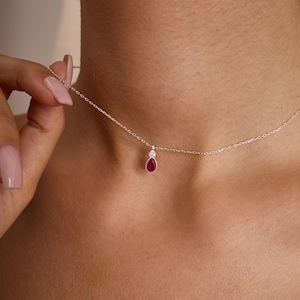 May include: A delicate silver necklace featuring a teardrop-shaped ruby pendant accented with a small, round diamond above it. The necklace is shown against a person's skin, highlighting the jewelry's fine details and elegant design.