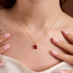 Puede incluir: Un collar dorado con un colgante rectangular de piedra preciosa rojo oscuro. El colgante cuelga de una cadena fina. El collar se muestra sobre un fondo claro.