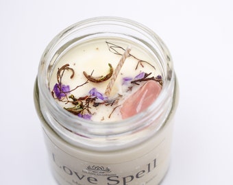 May include: A glass jar candle with a white wax base and a pink crystal embedded in the wax. The candle is topped with dried flowers and herbs. The label on the jar reads "Love Spell".