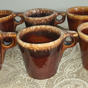 May include: A set of five ceramic mugs featuring a glossy dark brown glaze with a distinctive cream-colored drip pattern around the upper rim. Each mug has a sturdy, rounded handle and a wide, open mouth, showcasing a consistent design.
