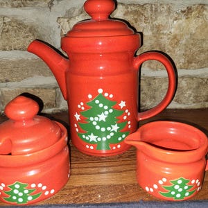 May include: A red ceramic Christmas tea set featuring a teapot, sugar bowl, and creamer. Each piece is adorned with a green Christmas tree design with white accents. The set is displayed on a wooden surface against a stone background.