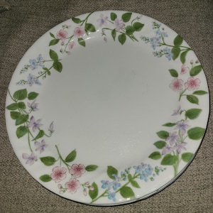 May include: White ceramic plates with a floral design. The plates feature a border of pink, purple, and blue flowers with green leaves. The plates are stacked on top of each other.