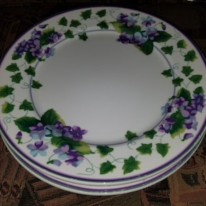 May include: A stack of white ceramic plates featuring a decorative border of purple violet flowers and green ivy leaves. The rim of each plate is accented with a thin purple line, framing the floral pattern that encircles the center of the dish.