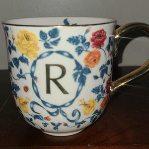 May include: White ceramic mug with a gold handle and rim, decorated with colourful roses and blue foliage. The letter "R" is centered within a blue ribbon design. The mug's floral pattern includes yellow, red, and orange roses.