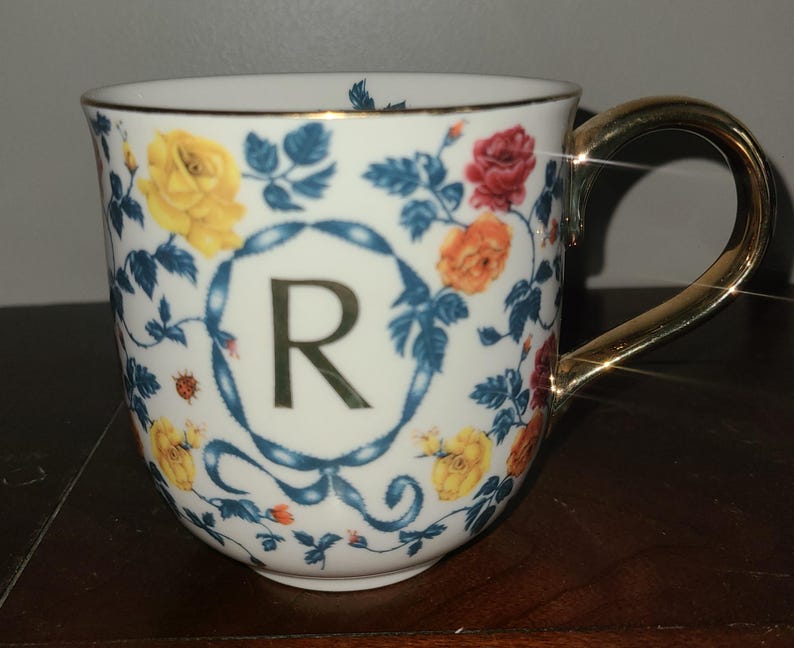 May include: White ceramic mug with a gold handle and rim, decorated with colourful roses and blue foliage. The letter "R" is centered within a blue ribbon design. The mug's floral pattern includes yellow, red, and orange roses.