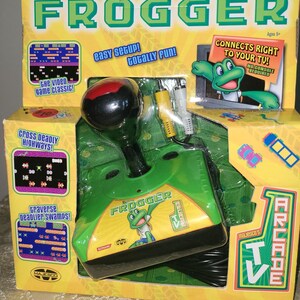 May include: A boxed Frogger arcade game for TV. The box is yellow and green with the game's logo and a frog character. The game controller is green with a black joystick and includes the text "Easy Setup!" and "Connects Right to Your TV!"