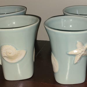 May include: Four light blue ceramic mugs with white seashell and starfish relief designs. Each mug has a curved handle and a slightly flared shape. The mugs are arranged on a dark brown surface.