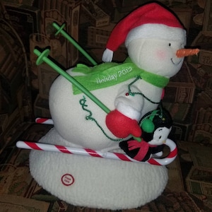 May include: A plush snowman wearing a red Santa hat and green scarf is skiing down a candy cane sled with a penguin. The snowman has a carrot nose and red cheeks. The penguin is wearing a green hat and a red scarf. The snowman's skis are green and the sled is white with red stripes. The snowman is holding a green ski pole in his right hand. The penguin is holding a green ski pole in its left flipper. The snowman's scarf has the text "Holiday 2012" embroidered on it. The snowman is sitting on a white base with the words "Press Here" printed on it.