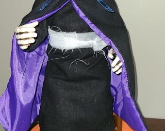 May include: A Halloween decoration of a skeleton figure wearing a black hooded robe with purple lining. The skeleton has a white skull with red eyes and is standing on a round orange and purple base.