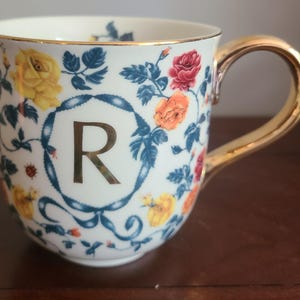 May include: A white ceramic mug featuring a gold handle and rim, decorated with roses and blue leaves. The letter 'R' is displayed in the centre, framed by a blue ribbon design. The mug is a decorative item.