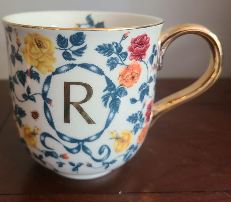 May include: A white ceramic mug featuring a gold handle and rim, decorated with roses and blue leaves. The letter 'R' is displayed in the centre, framed by a blue ribbon design. The mug is a decorative item.