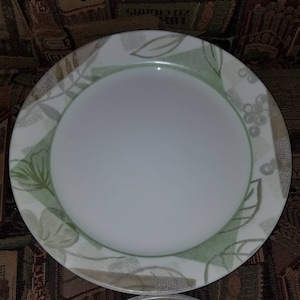 May include: A white ceramic dinner plate with a green and brown floral design. The plate has a white rim and a slightly raised edge.