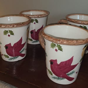 May include: A set of four white ceramic mugs featuring a painted design of red cardinal birds, green holly leaves, and red berries. Each mug has a textured, brown, branch-like rim and a matching handle, creating a rustic appearance.