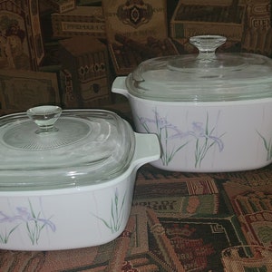 May include: Two matching white casserole dishes with glass lids. Each dish features a floral design of purple irises and green stems. The dishes have a square shape with rounded corners and a handle on each side.