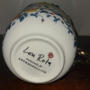 May include: White ceramic mug with a gold handle and floral design. The mug features a delicate pattern of blue leaves, orange and yellow flowers. The bottom of the mug has the text "Lou Rota exclusively for ANTHROPOLOGIE" and a ladybird illustration.