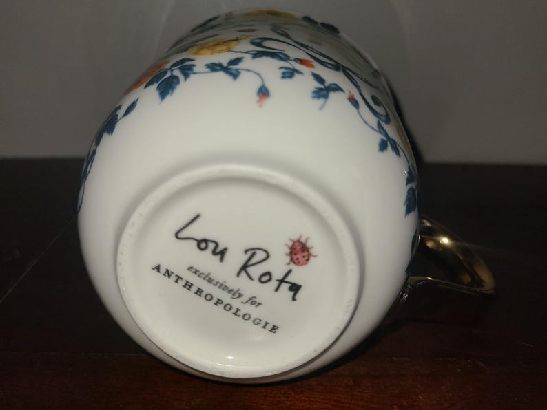May include: White ceramic mug with a gold handle and floral design. The mug features a delicate pattern of blue leaves, orange and yellow flowers. The bottom of the mug has the text "Lou Rota exclusively for ANTHROPOLOGIE" and a ladybird illustration.