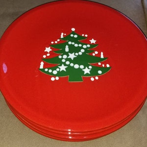May include: A stack of red plates with a green Christmas tree design. The tree is decorated with white stars, lights, and candles. The plates are round and have a glossy finish, perfect for holiday meals.