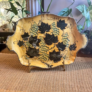 May include: A decorative tray with a scalloped edge features a hand-painted design of grapes and leaves. The tray is yellow with black and green accents. The tray is on a gold metal stand.