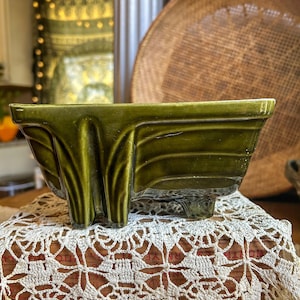 May include: A rectangular, olive green ceramic planter with a textured, Art Deco-inspired design. The planter sits on a white crocheted doily. A woven basket is in the background.