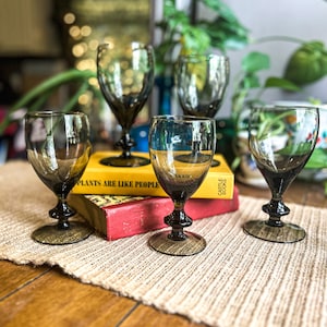 May include: Four clear glass wine glasses with a dark green tint, sitting on a woven placemat on a wooden table. The glasses are arranged in a semi-circle, with two glasses in the front and two glasses in the back. The glasses are resting on top of a stack of three books, the top book has the text "Castle Books" printed on the spine.