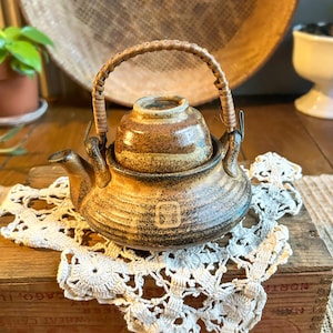 May include: A ceramic teapot with a woven handle and lid. The teapot is brown with darker brown and black accents. It sits on a white lace doily atop a wooden box. The teapot has a spout and a small square design on the front.