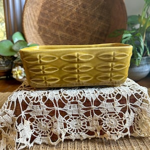 May include: A rectangular, yellow-green ceramic planter with a repeating oval and cross pattern. The planter sits on a vintage, cream-colored lace doily and a burlap surface. A woven basket and greenery are in the background.