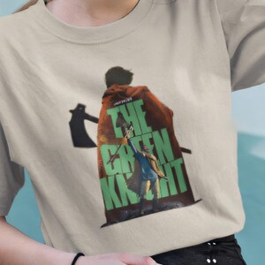 Op de afbeelding: Een beige t-shirt met een grafische print van een persoon die een bijl vasthoudt met de tekst "The Green Knight" in groene letters.