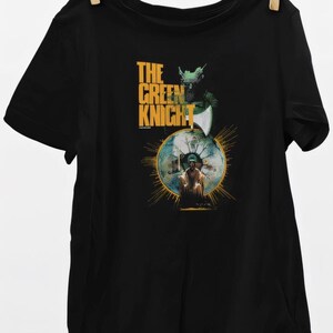 May include: Black t-shirt featuring a graphic from "The Green Knight." The design includes the text "THE GREEN KNIGHT" in gold, along with an illustration of a knight and a figure within a golden circle. The shirt is hanging from a clothesline.