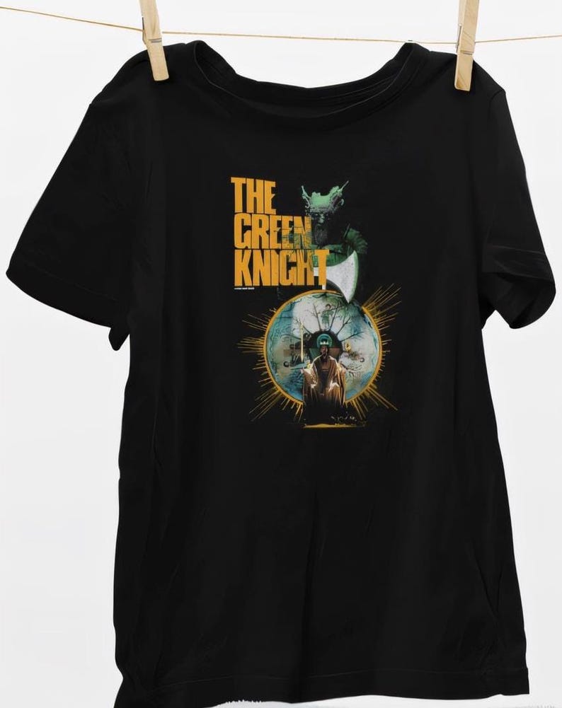 May include: Black t-shirt featuring a graphic from "The Green Knight." The design includes the text "THE GREEN KNIGHT" in gold, along with an illustration of a knight and a figure within a golden circle. The shirt is hanging from a clothesline.