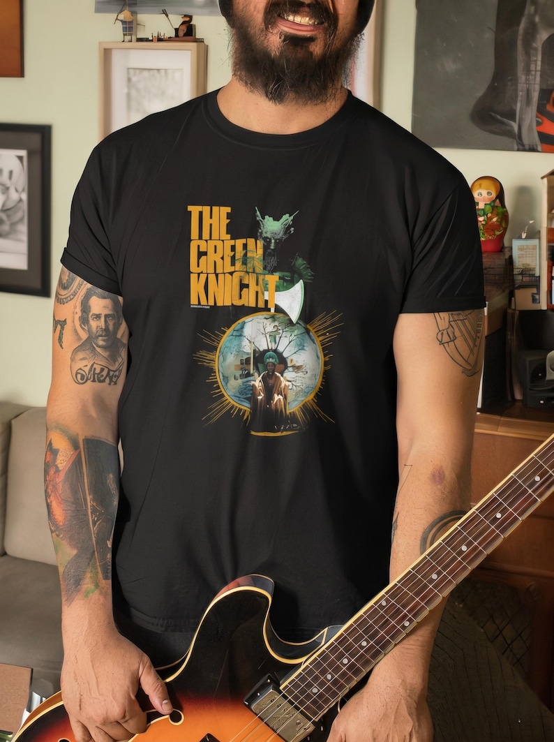 May include: Black t-shirt featuring a graphic from "The Green Knight" film. The design includes the film's title in yellow and green, along with an illustration of a knight and an axe. The shirt is made of a soft material.