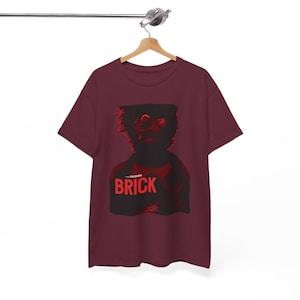 May include: A maroon t-shirt featuring a red and black graphic of a person's face and the word "BRICK" in red block letters. The shirt is hanging on a wooden hanger.