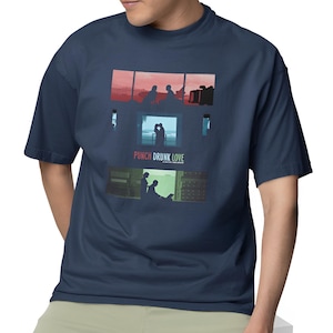 May include: Navy blue t-shirt featuring a graphic design with three rectangular panels. The panels depict silhouettes of people in various scenes, with the text "PUNCH DRUNK LOVE" below the middle panel. The shirt is a short-sleeved crew neck.