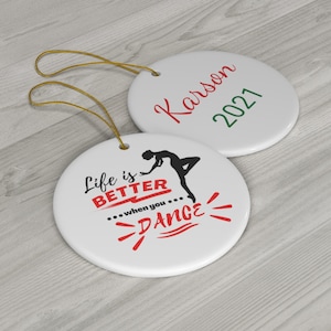 May include: Two round white ceramic ornaments with gold string. One ornament has a silhouette of a dancer with the text "Life is Better... when you... DANCE!" in red. The other ornament has the text "Karson 2021" in red and green.