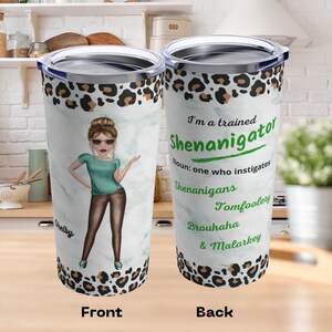 May include: A stainless steel tumbler with a white background and a leopard print design. The front features a cartoon illustration of a woman with brown hair and sunglasses. The back features the text "I'm a trained Shenanigator. Noun: one who instigates Shenanigans, Tomfoolery, Brouhaha & Malarkey."