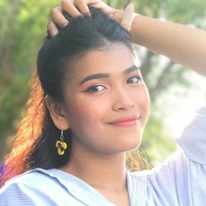 May include: A woman with long black hair is wearing a light blue button-down shirt with a white stripe pattern. She is wearing yellow flower-shaped earrings.