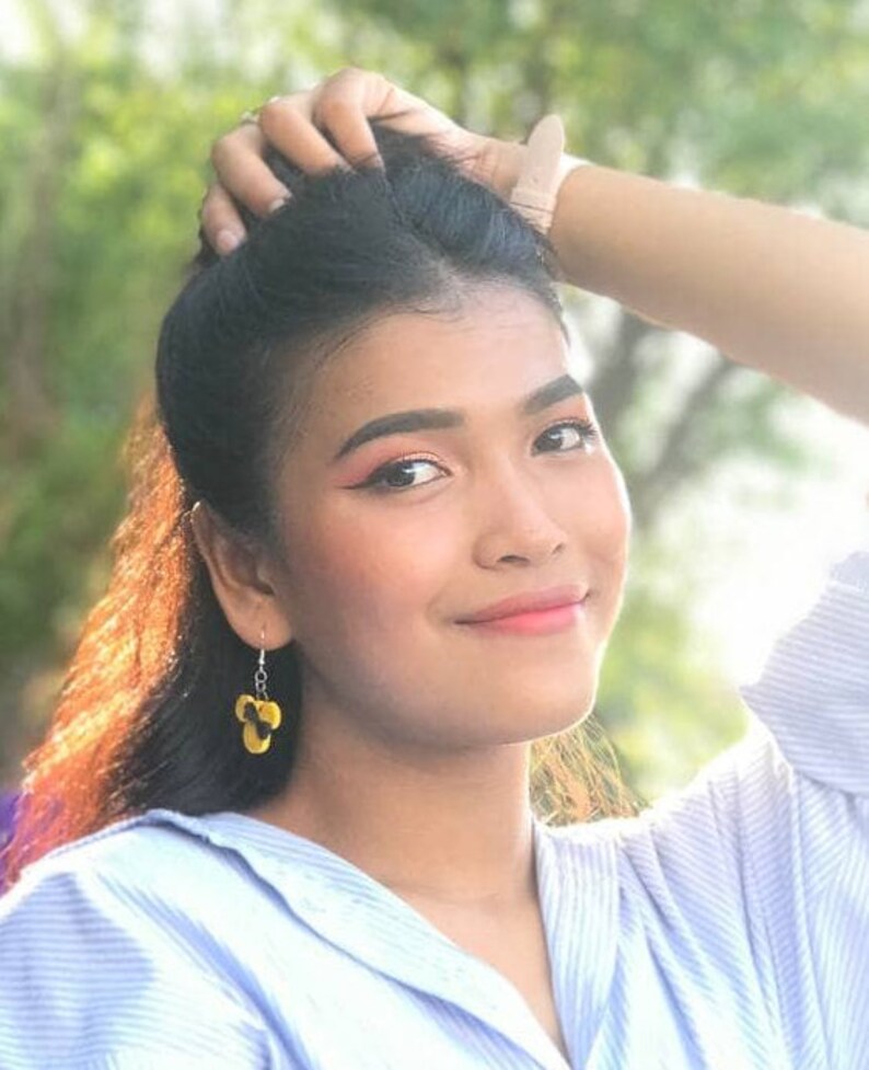 May include: A woman with long black hair is wearing a light blue button-down shirt with a white stripe pattern. She is wearing yellow flower-shaped earrings.