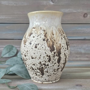Puede incluir: Un jarrón de cerámica con un diseño moteado. El jarrón tiene una parte superior de color crema que se desvanece en un patrón moteado de marrón y crema. El jarrón tiene una boca ancha y un cuerpo redondeado. El jarrón mide aproximadamente 20 cm de alto.