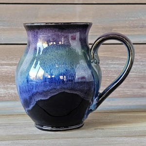 Può includere: Una tazza in ceramica fatta a mano con finitura lucida. La tazza presenta una sfumatura di colori, che passa da una base nera profonda a sfumature di blu, verde acqua e viola nella parte superiore. Il manico è in tinta. La tazza è progettata per bevande calde.