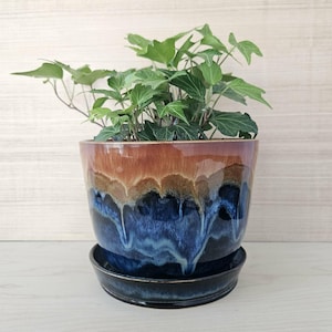 May include: A ceramic plant pot with a glossy finish, featuring a gradient of colors from brown to blue. The pot holds a green plant with multiple leaves. The pot sits on a matching saucer, creating a cohesive look for indoor gardening.
