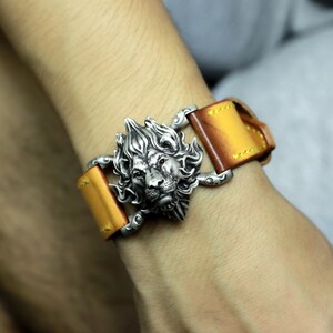 May include: A leather bracelet with a silver lion head buckle. The bracelet is a light brown color and has a distressed look.