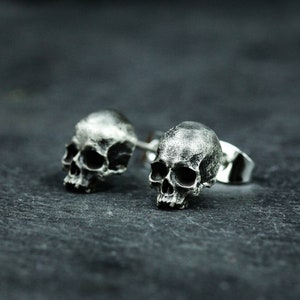 Puede incluir: Un par de pendientes de botón de calavera de plata. Las calaveras son detalladas y tienen un aspecto realista.